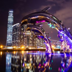 Dramatic cityscape at night featuring illuminated dolphin sculpture with reflections.